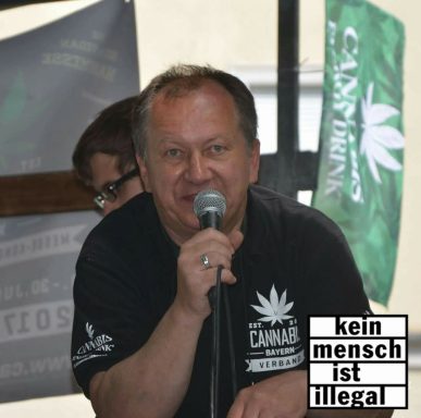 Mann spricht am Mikrofon, mit Cannabis-Flaggen im Hintergrund.